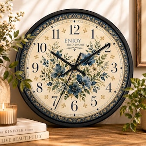 May include: A round wall clock with a black frame, featuring a cream-colored face with dark blue numerals, a floral pattern in shades of blue and green, and the text "ENJOY the Moment" printed in the center with black clock hands.