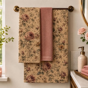 May include: Two rectangular bath towels featuring a beige background with a detailed floral pattern of pink roses and green foliage, paired with a solid dusty rose towel hanging on a dark metal towel bar in a bathroom setting.