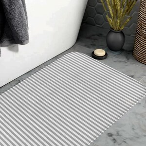 May include: A rectangular bath mat featuring a pattern of thin, alternating white and light gray stripes. The mat has rounded corners and a flat, uniform texture, positioned on a gray tiled floor in front of a white bathtub.