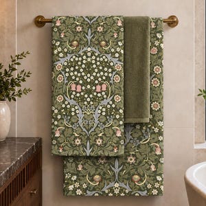 May include: Two bath towels hanging on a brass rod. One towel features a dense botanical pattern with green leaves, white flowers, and pink bell-shaped blossoms. The second towel is a solid, muted olive green color with a soft texture.