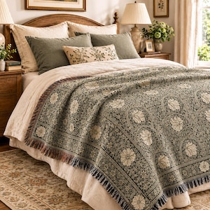 May include: A woven throw blanket featuring a dark green and cream floral pattern with repeating circular motifs and a fringed edge. The blanket is draped over a bed with light-colored bedding, olive green pillows, and a small rectangular accent pillow.