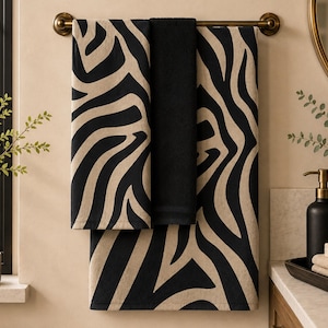 May include: A set of three rectangular bathroom towels hanging on a bronze-coloured metal bar. Two towels feature a bold black and beige zebra stripe pattern, while the central towel is a solid, deep black colour with a soft, plush texture.