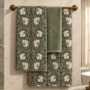 May include: Two bath towels hanging on a bronze-colored metal bar. One towel features a repeating pattern of white flowers, green leaves, and small yellow blossoms on a dark background. The second towel is a solid, muted sage green color.