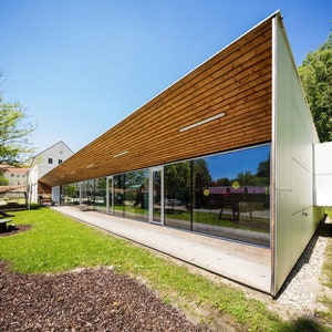 Op de afbeelding: Een modern, laag gebouw met een dak van houten panelen en grote ramen. Het gebouw is omgeven door groen gras en bomen.