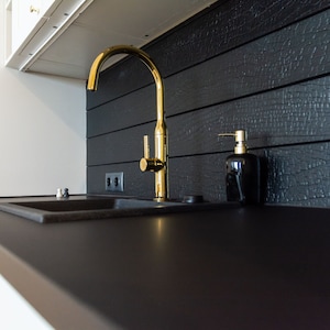 Könnte beinhalten: Ein goldener Küchenhahn mit einem einzigen Griff und einem hohen Auslauf ist auf einer schwarzen Arbeitsplatte montiert. Rechts vom Hahn befindet sich ein schwarzer Seifenspender. Die Arbeitsplatte befindet sich vor einer schwarzen Fliesenrückwand.