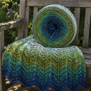 Könnte beinhalten: Ein großes, rundes Garnknäuel mit einem Farbverlauf von tiefem Marineblau bis zu hellem Limettengrün, das auf einer gestrickten Decke liegt, die denselben Ombré-Farbverlauf und ein sich wiederholendes Zickzack-Spitzenmuster aufweist.