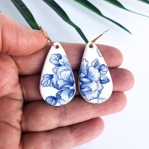 Op de afbeelding: Een paar traanvormige oorbellen met een witte achtergrond en een blauw bloemenpatroon. De oorbellen zijn gemaakt van keramiek en hebben gouden haakjes.