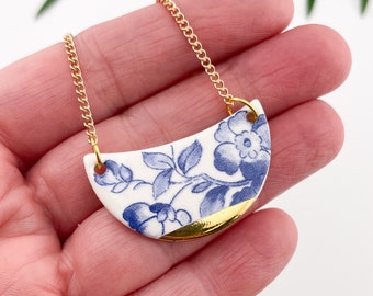 May include: A gold chain necklace with a blue and white porcelain pendant. The pendant is a half-circle shape with a floral design and a gold trim.