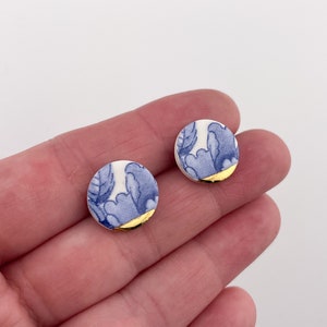 May include: A pair of round stud earrings. Each earring features a blue and white floral pattern with a gold-colored edge. The earrings are small and delicate, suitable for everyday wear.