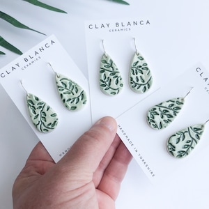 May include: A set of three pairs of green and white ceramic earrings with a leaf design. The earrings are handmade in Yorkshire and are packaged on white cards with the brand name "Clay Blanca Ceramics".