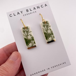 Op de afbeelding: Rechthoekige keramische oorbellen in crème met een groen botanisch patroon en gouden accenten. De oorbellen worden getoond op een witte kaart met de tekst "CLAY BLANCA CERAMICS" en "HANDMADE IN YORKSHIRE". Goudkleurige haken.
