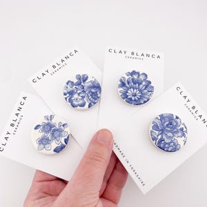 May include: Four round ceramic brooches with blue floral designs on white cards. The cards display the text "CLAY BLANCA CERAMICS" and "HANDMADE IN YORKSHIRE". The brooches are held in a hand.