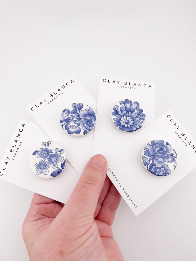 May include: Four round ceramic brooches with blue floral designs on white cards. The cards display the text "CLAY BLANCA CERAMICS" and "HANDMADE IN YORKSHIRE". The brooches are held in a hand.