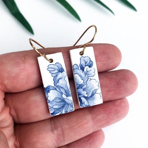 Op de afbeelding: Een paar blauwe en witte porseleinen oorbellen met een bloemenmotief. De oorbellen zijn rechthoekig en hebben gouden haakjes.