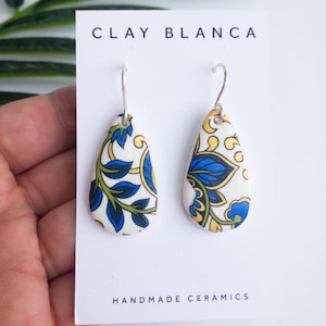 Op de afbeelding: Een paar witte keramische oorhangers met een blauw en geel bloemenpatroon. De oorhangers bevinden zich op een witte kaart met de tekst "CLAY BLANCA HANDMADE CERAMICS".
