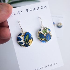 Puede incluir: Un par de pendientes de cerámica hechos a mano con un patrón floral azul y blanco. Los pendientes están en una tarjeta blanca con el texto "Clay Blanca Handmade Ceramics".
