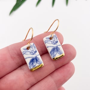 Könnte beinhalten: Ein Paar blaue und weiße Porzellanohrringe mit goldenen Verzierungen. Die Ohrringe sind rechteckig und haben ein Blumenmuster.