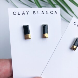 May include: A pair of black and gold rectangular ceramic earrings. The earrings are displayed on a white card with the text "CLAY BLANCA" printed on it.