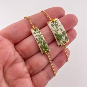 Könnte beinhalten: Ein Paar rechteckige Ohrringe mit einem floralen Design in Grün und Weiß. Die Ohrringe sind mit einer goldenen Kette verbunden. Die Ränder der Ohrringe sind mit Gold verziert.