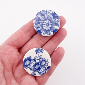 May include: Two round, white ceramic buttons with blue floral designs. One button features a large daisy-like flower, while the other displays a cluster of smaller flowers. The buttons are held in a hand, showcasing their size and detail.