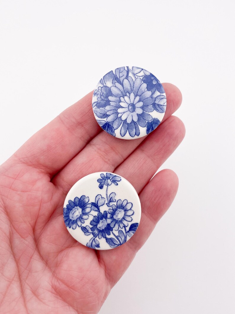 May include: Two round, white ceramic buttons with blue floral designs. One button features a large daisy-like flower, while the other displays a cluster of smaller flowers. The buttons are held in a hand, showcasing their size and detail.
