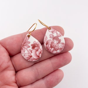 May include: A pair of dangle earrings with a teardrop-shaped design. The earrings are made of white ceramic with a red floral pattern. The earrings have gold-colored hooks.