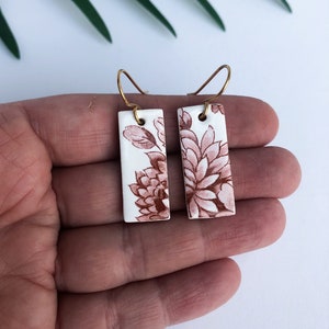 May include: A pair of white ceramic earrings with a brown floral pattern. The earrings are rectangular in shape and have a gold wire hook.