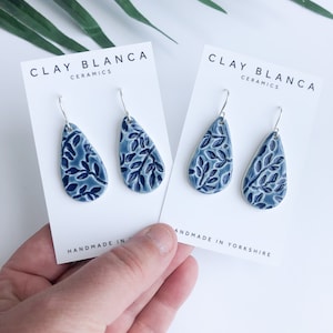 Puede incluir: Dos pares de pendientes de cerámica azul con forma de lágrima y diseño de hojas, expuestos sobre tarjetas blancas. Las tarjetas indican "CLAY BLANCA CERAMICS" y "HANDMADE IN YORKSHIRE". Los pendientes tienen ganchos plateados.