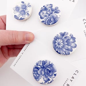 May include: Four ceramic brooches with blue floral designs on a white background. Each brooch is round and features a different floral pattern. The cards behind the brooches have the text "CLAY BLANCA CERAMICS".