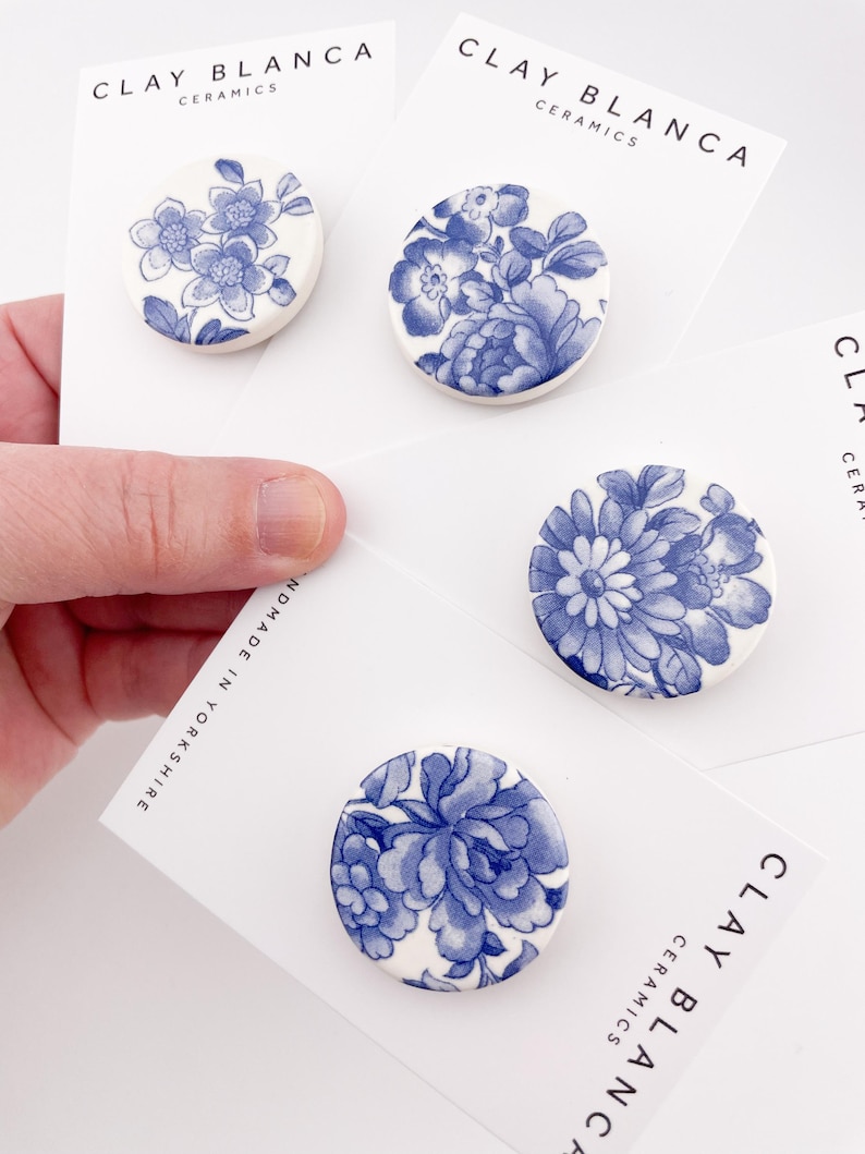 May include: Four ceramic brooches with blue floral designs on a white background. Each brooch is round and features a different floral pattern. The cards behind the brooches have the text "CLAY BLANCA CERAMICS".