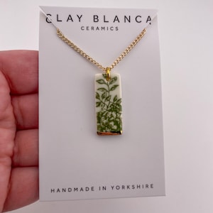 Op de afbeelding: Een ketting met een gouden ketting en een rechthoekige hanger. De witte hanger heeft een groen bloemmotief en een gouden rand. De kaart leest "CLAY BLANCA CERAMICS" en "HANDMADE IN YORKSHIRE."