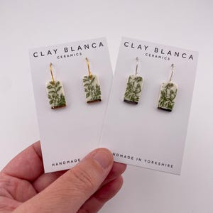 May include: Two pairs of rectangular ceramic earrings with a botanical design, displayed on white cards. The earrings have gold-coloured trim and green detailing. The cards read "CLAY BLANCA CERAMICS" and "HANDMADE IN YORKSHIRE."