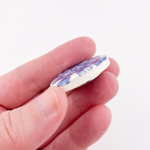 May include: A round, white ceramic pendant with a floral design in shades of purple and blue. The pendant is held in a hand against a white background. The floral pattern is detailed and covers the surface of the pendant.