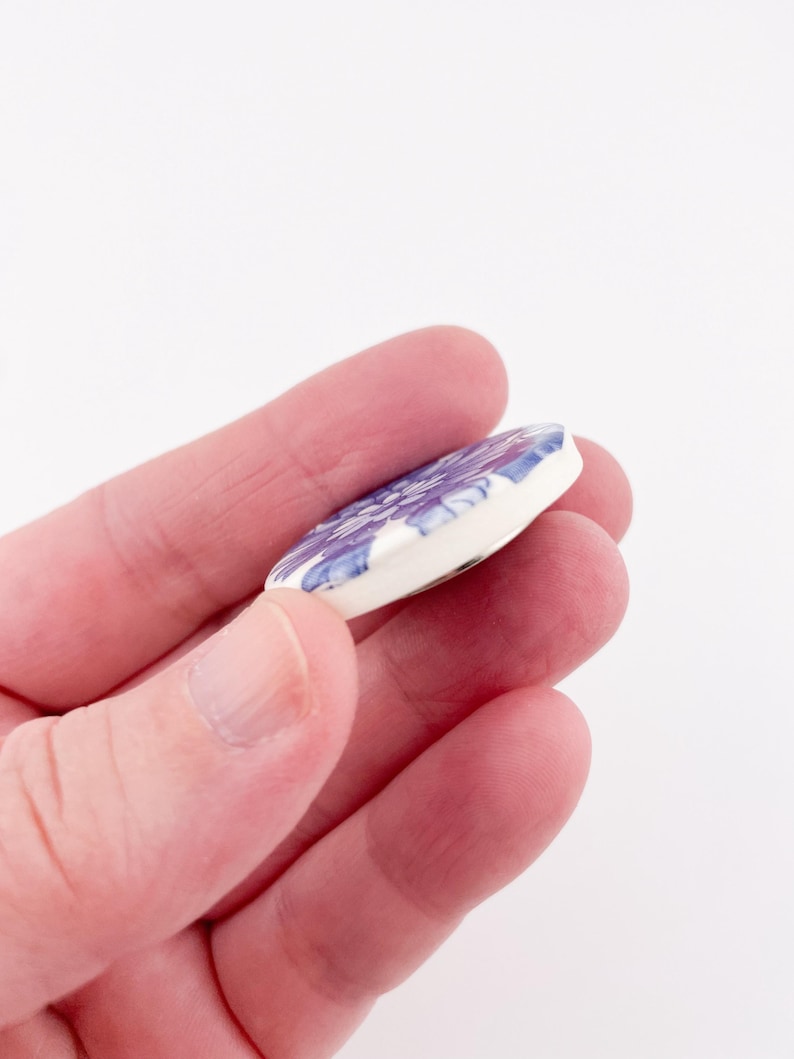 May include: A round, white ceramic pendant with a floral design in shades of purple and blue. The pendant is held in a hand against a white background. The floral pattern is detailed and covers the surface of the pendant.