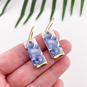 Op de afbeelding: Een paar blauwe en witte porseleinen oorbellen met gouden accenten. De oorbellen zijn rechthoekig en hebben een bloemenmotief.