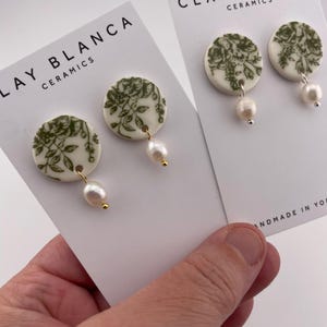 May include: Round white ceramic earrings with a green floral pattern and a dangling pearl. The earrings are displayed on cards that read "CLAY BLANCA CERAMICS". The earrings are held in a hand.