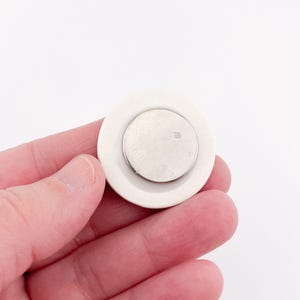 May include: A white, circular object with a smaller, silver-colored disc in the center. The object is held in a hand against a white background. The design is simple and minimalist, with a focus on the contrast between the white and silver elements.