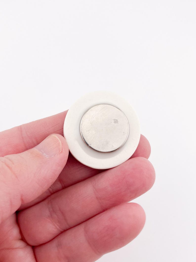 May include: A white, circular object with a smaller, silver-colored disc in the center. The object is held in a hand against a white background. The design is simple and minimalist, with a focus on the contrast between the white and silver elements.
