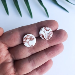 Op de afbeelding: Een paar witte keramische oorbel studs met een bruin bloemenpatroon.
