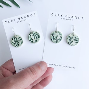 May include: A pair of green ceramic earrings with a leafy design. The earrings are handmade in Yorkshire and are sold by Clay Blanca Ceramics.