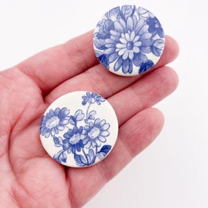 May include: Two round, white ceramic pieces with blue floral designs. One features a large flower, the other a cluster of smaller blooms. The pieces are held in a hand, showcasing the detailed blue and white patterns.