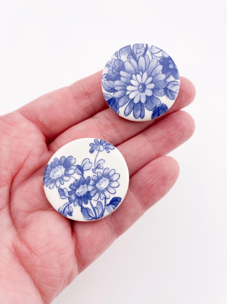 May include: Two round, white ceramic pieces with blue floral designs. One features a large flower, the other a cluster of smaller blooms. The pieces are held in a hand, showcasing the detailed blue and white patterns.