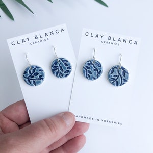 May include: A pair of blue and white ceramic earrings with a leaf design. The earrings are on a white card with the text "CLAY BLANCA CERAMICS" and "HANDMADE IN YORKSHIRE".