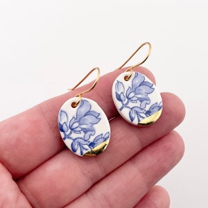 Puede incluir: Un par de pendientes de cerámica blanca y dorada con un diseño floral azul. Los pendientes son de forma ovalada y tienen un borde dorado.
