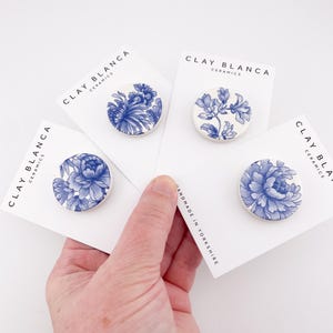 May include: Four ceramic brooches with blue floral designs on white cards. The cards are held in a hand. The brooches have different floral patterns. The cards read "CLAY BLANCA CERAMICS" and "HANDMADE IN YORKSHIRE".