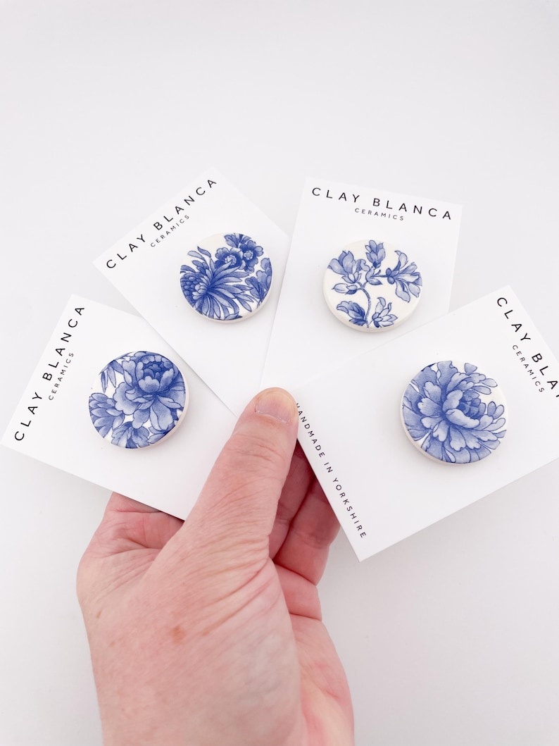 May include: Four ceramic brooches with blue floral designs on white cards. The cards are held in a hand. The brooches have different floral patterns. The cards read "CLAY BLANCA CERAMICS" and "HANDMADE IN YORKSHIRE".