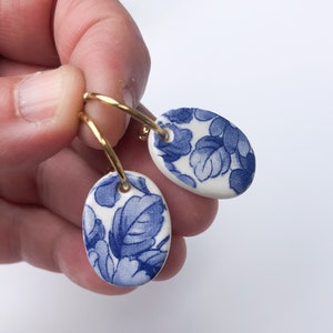 Puede incluir: Pendientes ovalados con un diseño floral azul sobre fondo blanco, colgados de aros dorados. El diseño presenta una combinación de colores azul y blanco clásica, que recuerda a la porcelana.
