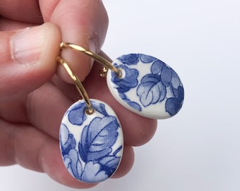 May include: Oval-shaped earrings with a blue floral pattern on a white background. The earrings are suspended from gold-colored hoops. The design features a classic blue and white color scheme, reminiscent of porcelain.