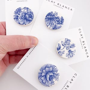 May include: Four ceramic brooches with blue floral designs on white cards. The cards display the text "CLAY BLANCA CERAMICS" and "HANDMADE IN YORKSHIRE". The brooches feature various blue floral patterns.