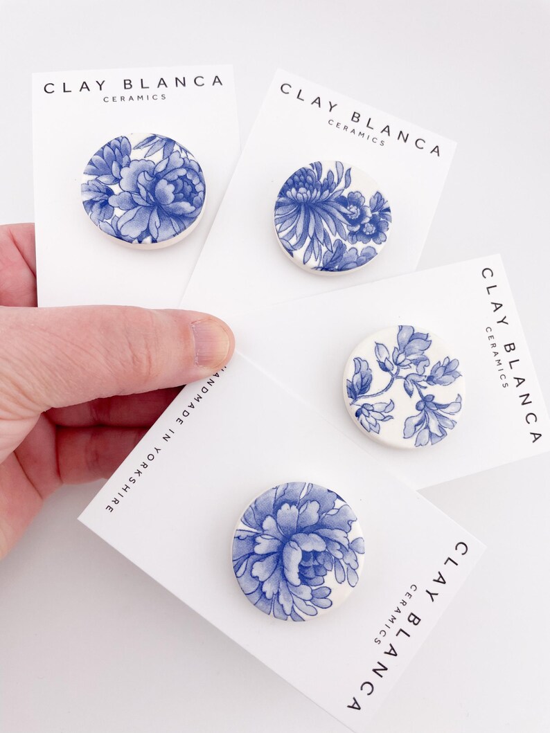 May include: Four ceramic brooches with blue floral designs on white cards. The cards display the text "CLAY BLANCA CERAMICS" and "HANDMADE IN YORKSHIRE". The brooches feature various blue floral patterns.
